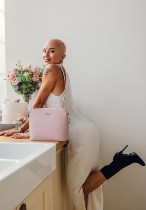 Pink crossbody bag with textured finish, silver hardware, and adjustable strap, placed on a wooden surface with floral arrangement in background.