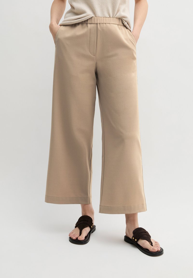 Person trägt beige weite Hosen mit elastischem Bund und schwarze Sandalen mit gekreuzten Riemen, steht mit den Händen in den Taschen.