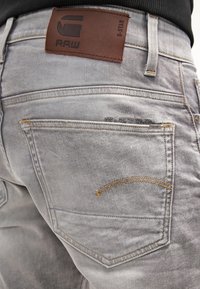 Szare dżinsy typu denim z skórzaną naszywką z napisem "RAW". Posiadają wygięty żółty szew na tylnej kieszeni i pięciokieszonkowy design.