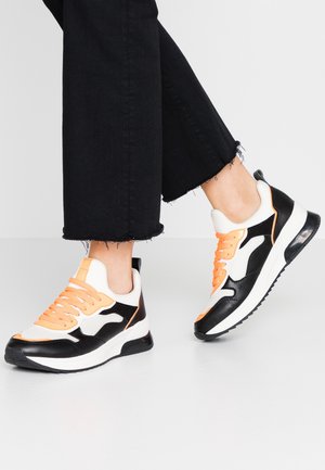 Person trägt schwarze kurze Hosen und schwarze, weiße und orangefarbene Sneakers mit dicken weißen Sohlen und steht auf einem weißen Boden.