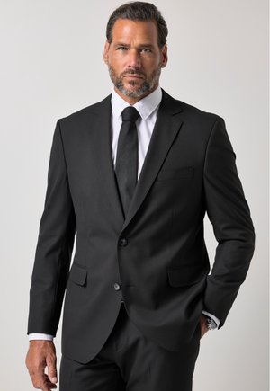 Hombre con pelo corto y barba, vestido con un traje negro, camisa blanca y corbata negra, de pie con una mano en el bolsillo sobre un fondo liso.