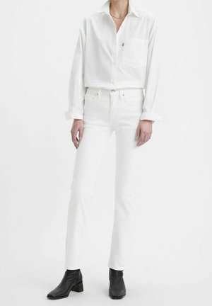 Personne portant une chemise blanche à manches longues et un jean blanc, debout, associée à des bottines noires sur un fond blanc uni.