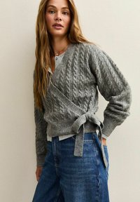 Maglione grigio lavorato a maglia con design avvolgente, caratterizzato da lavorazione a treccia e dettaglio in vita annodato, abbinato a jeans blu.