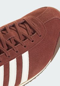 Κόκκινο αθλητικό παπούτσι από σουέτ με λευκές ζιγκ-ζαγκ ρίγες και υφασμένα κορδόνια. Διαθέτει στρογγυλεμένη μύτη και ελαφρώς καφέ εξωτερική σόλα.