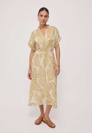 Vrouw die een beige midijurk draagt met gele bladpatronen en korte mouwen, gecombineerd met bruine open-toe sandalen en zilveren creolen.