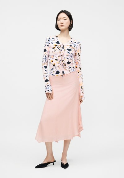 Femme portant un haut à manches longues rose à motifs floraux et cœurs, une jupe midi rose clair et des escarpins noirs à talons petits et pointus, debout devant un fond blanc.