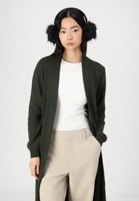 Lang, mørkegrønn strikkekardigan med ribbestruktur, brukt over en hvit topp, kombinert med beige vidde bukser, og komplettert med svarte pelskåper.