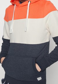 Felpa con cappuccio caratterizzata da blocchi di colore arancione, bianco e blu navy. Realizzata in cotone misto, presenta una tasca frontale a marsupio e chiusura con cordino.