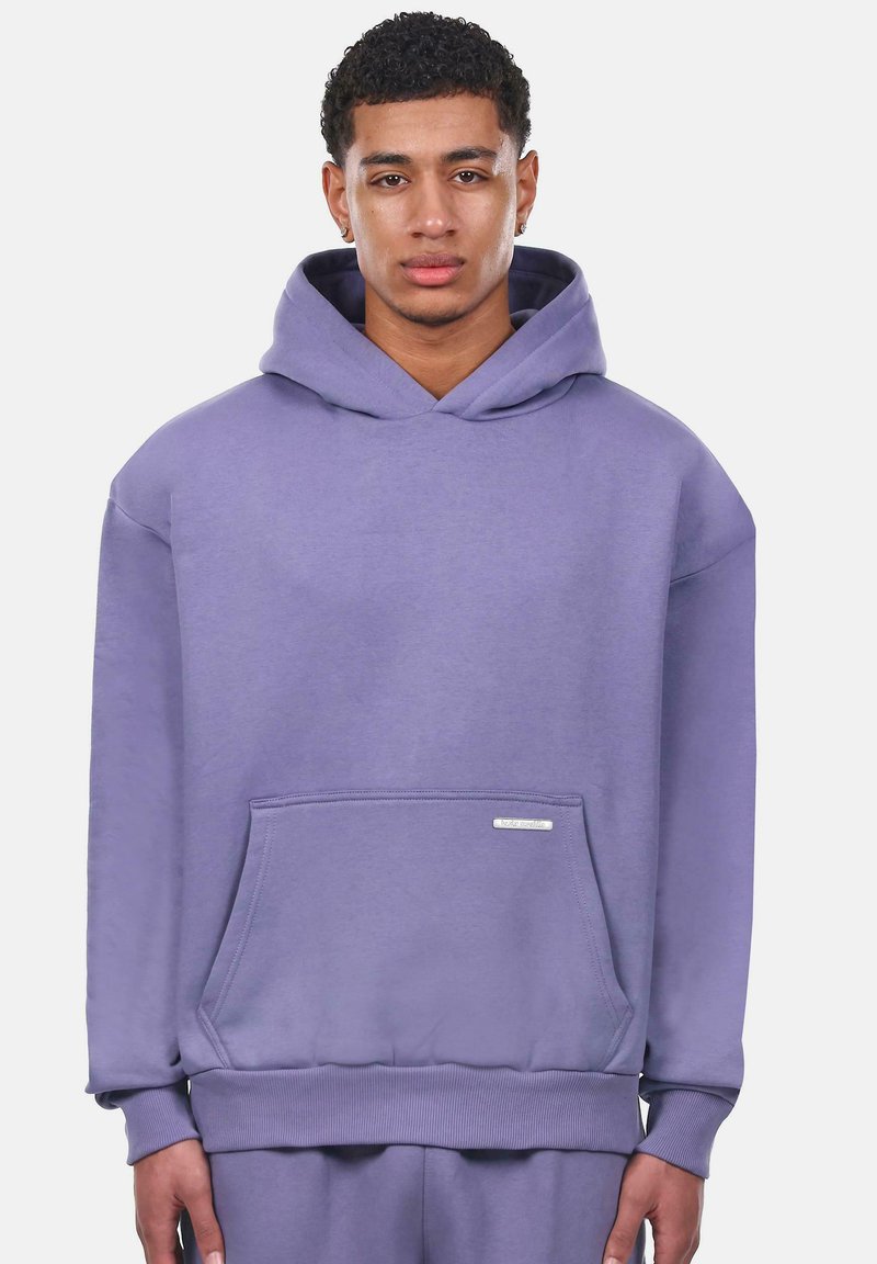 Lila Kapuzenpullover aus weichem Stoff, mit einer Fronttasche, gerippten Bündchen und einem kleinen Markenetikett auf der Tasche.