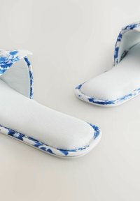 Tekstiilipohja varrettomat tossut, joissa on sininen kukkakuosi valkoisella taustalla, pehmeällä pehmustetulla pohjalla ja avoimella takatunnalla helppoa pukemista varten.