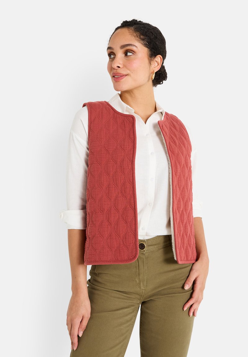 Gilet matelassé en rose poussiéreux, présentant un motif ondulé texturé et un design à ouverture frontale. Porté sur une chemise blanche, associé à un pantalon vert olive.
