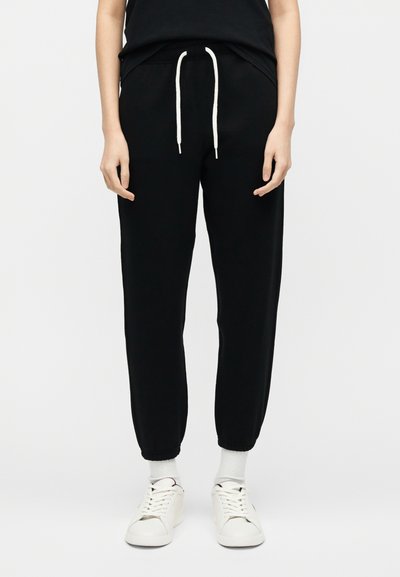 Άτομο που φοράει μαύρο παντελόνι τζόγκινγκ με λευκά κορδόνια, λευκές κάλτσες και λευκά αθλητικά παπούτσια, στέκεται μπροστά σε ένα απλό φωτεινό φόντο.