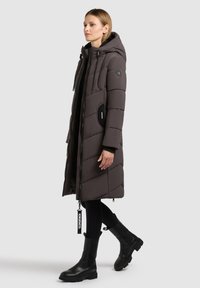 Dunkelgraue Steppjacke mit Kapuze, ausgestattet mit Zickzacknähten und schwarzen Akzenten, kombiniert mit schwarzen Knöchelstiefeln und Leggings. Reißverschluss.