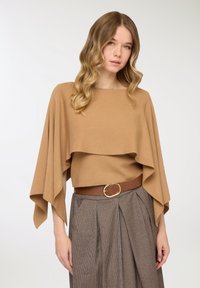 Top a strati color tan realizzato con materiale morbido, caratterizzato da un collo a barchetta e maniche ampie, abbinato a una cintura marrone su una gonna a quadri.