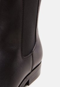 Gros plan d'une bottine en cuir noir avec panneau latéral élastique et petit talon empilé sur un fond clair uni.