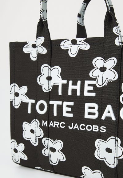 Čierna textilná taška s bielymi kvetinovými vzormi, ktorá má výrazný biely nápis "THE TOTE BAG" a "MARC JACOBS". Obsahuje pevné úchyty.