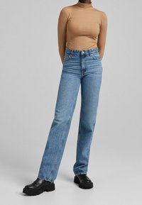 Person som bär en åtsittande ribbad långärmad topp i beige, högmidjade blå jeans och svarta plattformsskor, stående mot en enkel grå bakgrund.