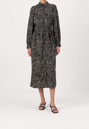 Kobieta ubrana w czarną sukienkę midi z białymi wzorami paisley, długimi rękawami, wiązanym w pasie i czarnymi sandałami na ramiączkach.