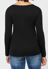 Woman with curly hair wearing a fitted black long-sleeve top and blue denim jeans, shown from the back against a plain background.