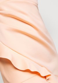 Hedvábná látka v broskvové barvě s jemným splývavým efektem, zdobená vlnitým volánkem podél okraje, vytvářející hladkou texturu a elegantní línii.