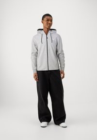 Sudadera gris con cremallera y capucha, bolsillos frontales y detalles en negro, combinada con pantalones holgados negros y zapatillas blancas.