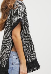 Poncho tricoté en noir et blanc avec des motifs géométriques, doté de manches courtes et de détails en frange le long de l'ourlet.