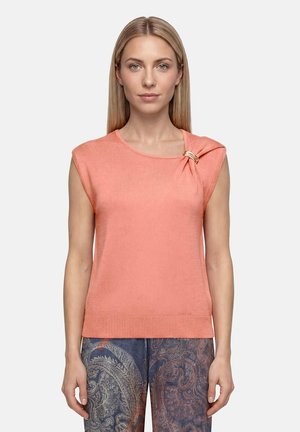 Femme aux cheveux blonds raides porte un haut en tricot corail sans manches avec un nœud décoratif et un pantalon bleu à motif cachemire, debout sur fond uni.