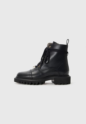 Bota de combate de cuero negro con suela gruesa de tacos, cordones en la parte delantera, ojetes dorados y tirador trasero sobre un fondo liso gris claro.