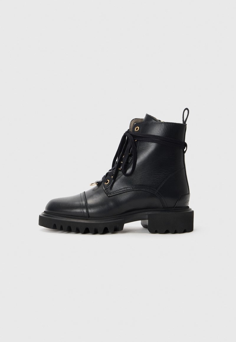 Botte de combat en cuir noir avec une semelle épaisse à crampons, un laçage frontal, des œillets dorés et une languette arrière, sur un fond uni gris clair.