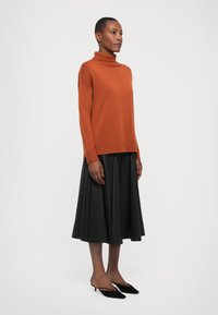 Rustfarget turtleneck genser med ribbestrikkede mansjetter, kombinert med et svart plisserte skjørt og svarte spisse mules på en nøytral bakgrunn.