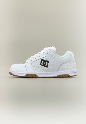 Chaussure de skate de la marque DC blanche avec logo noir sur le côté, lacets blancs et semelle en caoutchouc marron clair, sur un fond neutre.