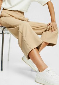 Personne assise sur une chaise en métal portant un pantalon beige large et court, des baskets blanches et un haut blanc à manches courtes.