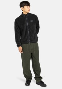 Giacca in pile nera con colletto alto, zip frontale e logo ricamato; abbinata a pantaloni larghi verde oliva e sneakers nere.
