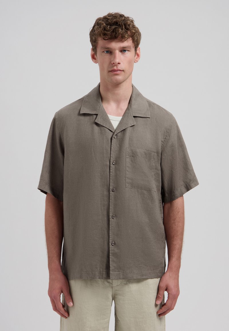 Homme debout face à l'avant, portant une chemise grise à manches courtes boutonnée sur un maillot blanc et un pantalon beige clair sur un fond uni.