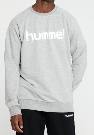 Homme portant un sweat-shirt gris à manches longues hummel et un pantalon noir hummel, debout devant un fond clair uni.