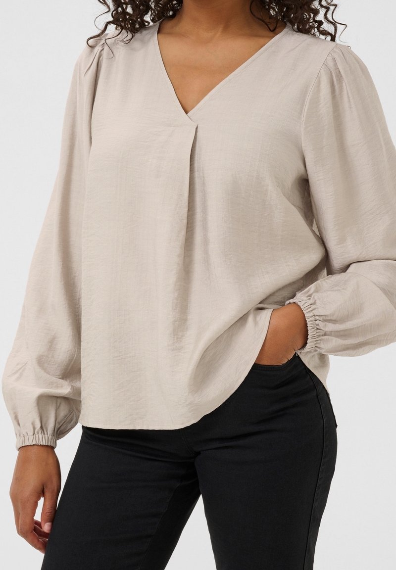 Blouse beige avec un col en V, un pli à l'avant et des poignets élastiques. Confectionnée dans un tissu léger à texture. Associée à un pantalon noir.
