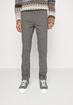 Osoba nosząca szare spodnie typu slim fit, szaro-białe trampki oraz wielokolorowy sweter w wzory stojąca na białym tle.