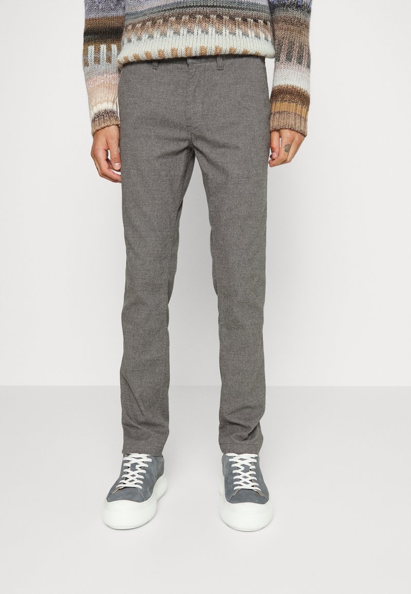 Personne portant un pantalon slim gris, des baskets grises et blanches, et un pull à motifs multicolores, debout devant un fond blanc.