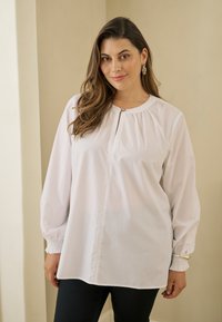 Femme aux longs cheveux bruns portant un chemisier blanc à manches longues avec des boutons et un pantalon noir, debout à l'intérieur contre des murs beiges.