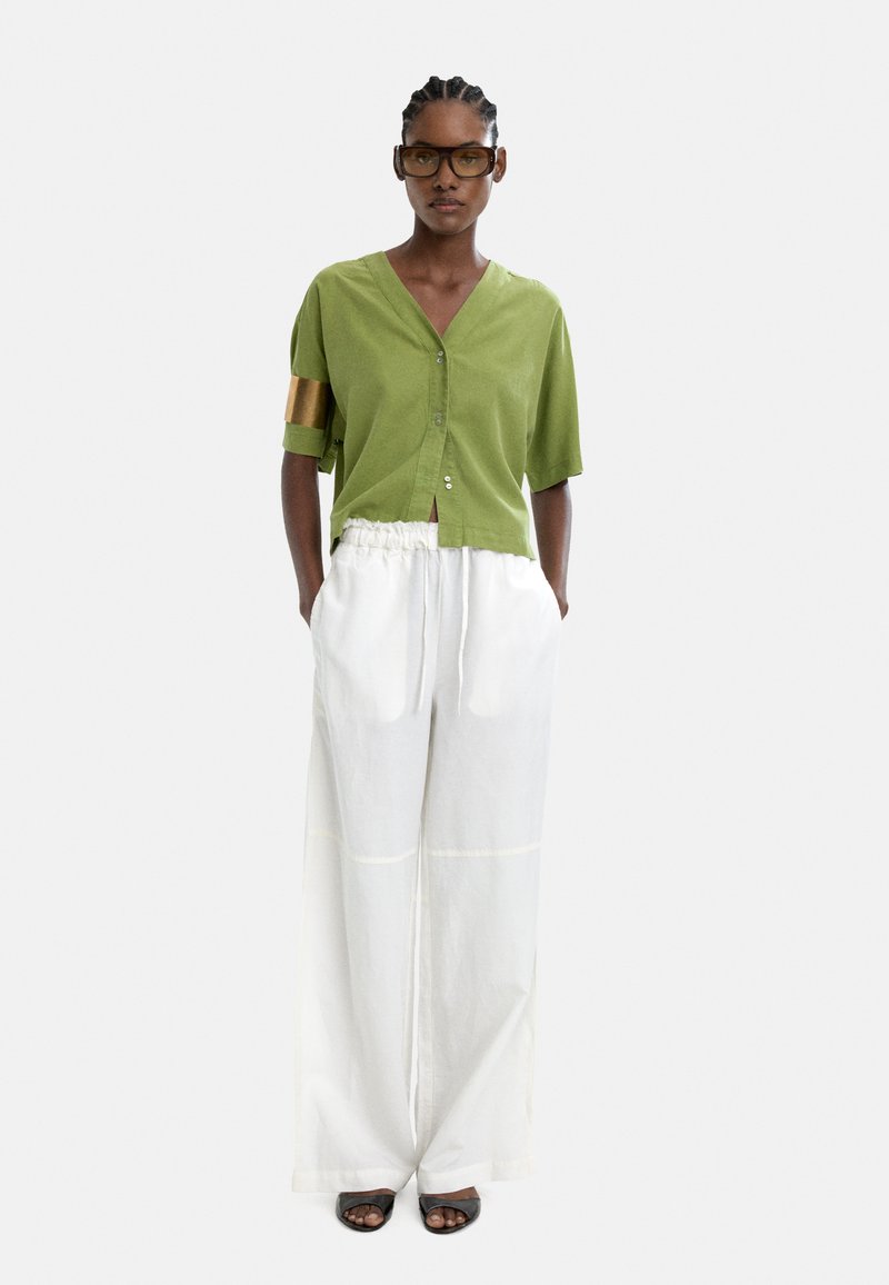 Zielona krótka koszula z guzikami i krótkim rękawem, z złotym akcentem; w komplecie z luźnymi białymi spodniami. Czarne sandały na odsłonięte palce.