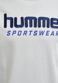 Jasnoszary t-shirt z wyraźnym, granatowym haftowanym napisem „humme” oraz „SPORTSWEAR” w czcionce bezszeryfowej. Miękka, teksturowana tkanina.