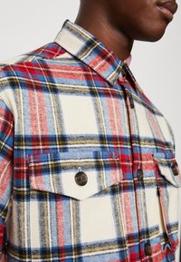Koszula flanelowa w kratę w kolorze kremowym, czerwonym, niebieskim i żółtym. Posiada wywinięty kołnierz, dwie kieszenie na piersi oraz zapięcie na guziki. Miękka tekstura.