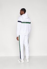 Fato de treino branco com capuz, apresentando detalhes em verde e azul-marinho. O casaco tem um logótipo nas costas, enquanto as calças têm riscas laterais.