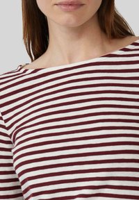 Vrouw in een wit en bordeauxrood horizontaal gestreept bootneck shirt, dat de nek, schouders en bovenborst toont.