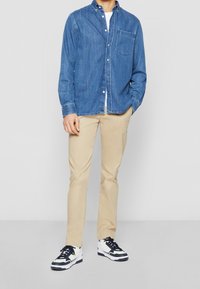Chemise en denim bleu foncé avec col boutonné, associée à un chino beige et des baskets noir et blanc. Comprend une poche poitrine et des boutons.