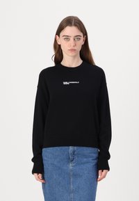 Črni pleteni pulover z dolgimi rokavi, rebrastimi manšetami in obrobo. Ima belo logotipno besedilo na prsih. Nosi se z jeans krilom.