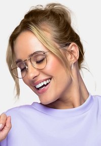 Runda metallglasögon med tunna bågar. Modellen bär en ljuslila topp, har en låg knut och visar ett subtilt leende. Ljus bakgrund.