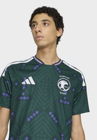 Green soccer jersey with purple geometric patterns, white Adidas logo and accents, crew neck design, short sleeves, and distinct Saudi emblem.
