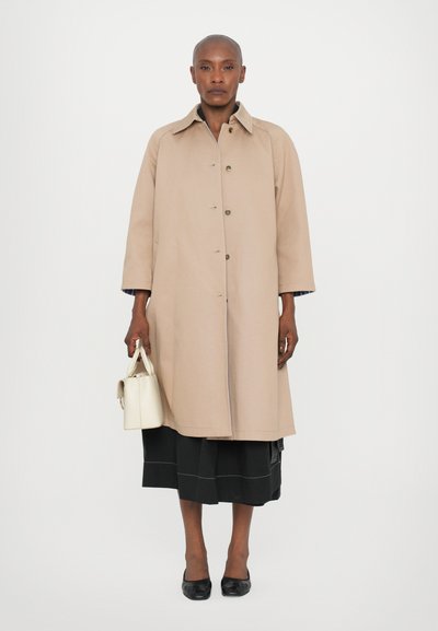 Cappotto trench beige con colletto e chiusura a bottoni, dotato di maniche lunghe e vestibilità rilassata. Un paio di scarpe nere e una piccola borsa bianca.