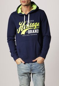 Marineblaue Kapuzenjacke mit grünem Innenfutter, ausgestattet mit einer Front-Kängurutasche und weißen Kordeln. "Heritage EDC BRAND"-Logo in Gelb.